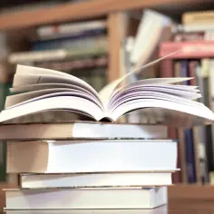 Otwarta książka na stosie innych, w tle regały pełne literatury, w tym "Zbrodnia i kara" gatunek.