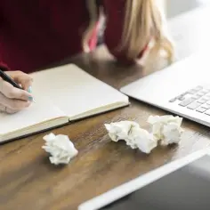Kobieta pisze w notatniku, szukając inspiracji, jak napisać recenzję. Obok leży laptop i pogięte kartki.