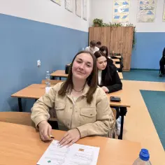Uśmiechnięta uczennica w technikum, kiedy jest półmetek. W tle inni uczniowie przy ławkach, na ścianach tablice z matematyki.