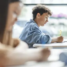 Chłopiec z kręconymi włosami pisze coś na kartce, obok laptop. Uczy się do egzaminu, który zdecyduje o jego **przejściu warunkowym w szkole średniej**.