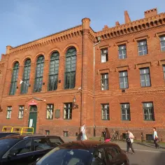 Ceglana kamienica z zielonymi oknami, gdzie w wieku 19 lat kończy się liceum. Ludzie idą chodnikiem.