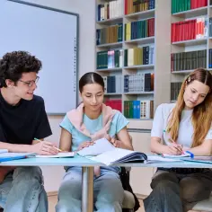 Trzech uczniów przy biurku w bibliotece, uczących się do egzaminów. Zastanawiają się, ile jest klas w technikum.