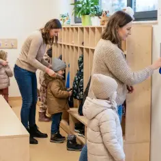 Rodzice przyprowadzają dzieci do przedszkola. W szatni opiekunowie pomagają maluchom się przebrać.