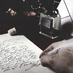 Dłoń trzyma pióro nad papierem z odręcznym pismem. Obok leży stempel. Czy to początek jakiejś fraszki?