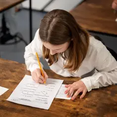 Dziewczynka w białej koszuli pisze coś ołówkiem na kartce z zadaniami matematycznymi.