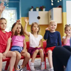 Dzieci na czerwonych krzesłach podnoszą ręce, jakby odpowiadały na pytanie, czy zerówka jest obowiązkowa.