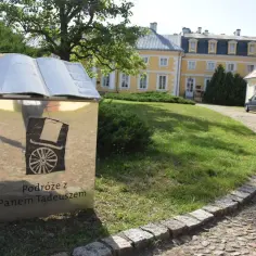 Obok dworku w Soplicowie, na tle żółtego budynku, stoi metalowa księga z napisem "Podróże z Panem Tadeuszem".