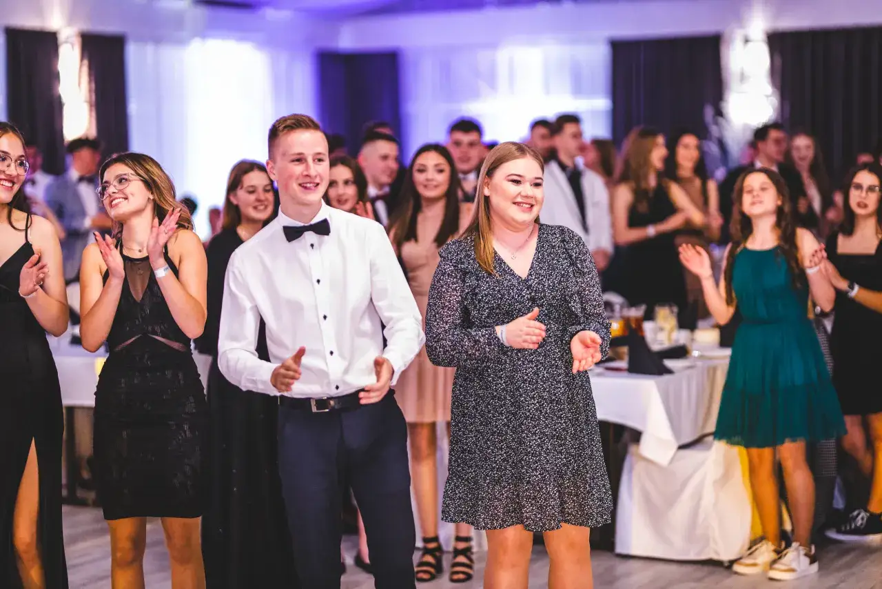 Uczniowie technikum świętują półmetek. Młodzi ludzie w eleganckich strojach bawią się na sali, w tle widać innych uczestników imprezy.